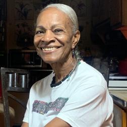 Caroline, an older African American woman with grey hair, sits at her kitchen table.