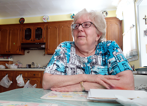 Jackie, Citymeals recipient, an older woman with white hair and glasses wearing a blue shirt, sits at her kitchen table at her home in Maspeth, Queens.