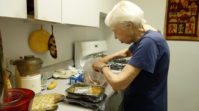 Deborah, an older New Yorker with white hair and glasses, prepares a Citymeals meal in her apartment.