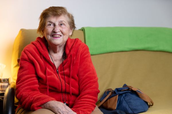 An older woman in a red shirt smiling and sitting on a couch.