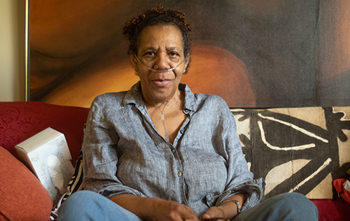 Elizabeth, an older African-American woman in a blue shirt and a portable oxygen machine, sits on her living room couch. 