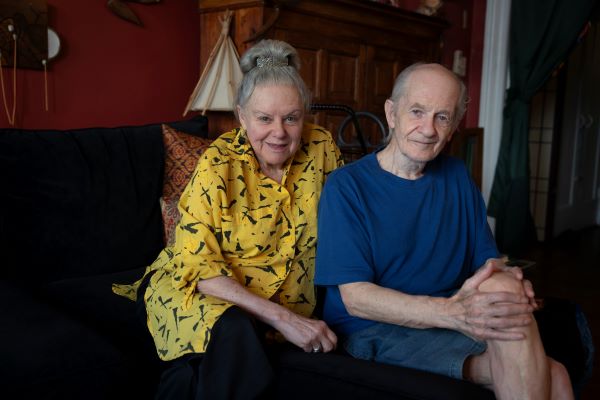 An older couple sit on the couch together, looking at the camera.