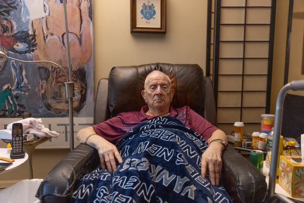 An older man, Lenny, sits in a leather recliner, surrounded by pill bottles.
