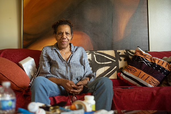 Elizabeth, an older African-American woman in a blue shirt and a portable oxygen machine, sits on her living room couch. 