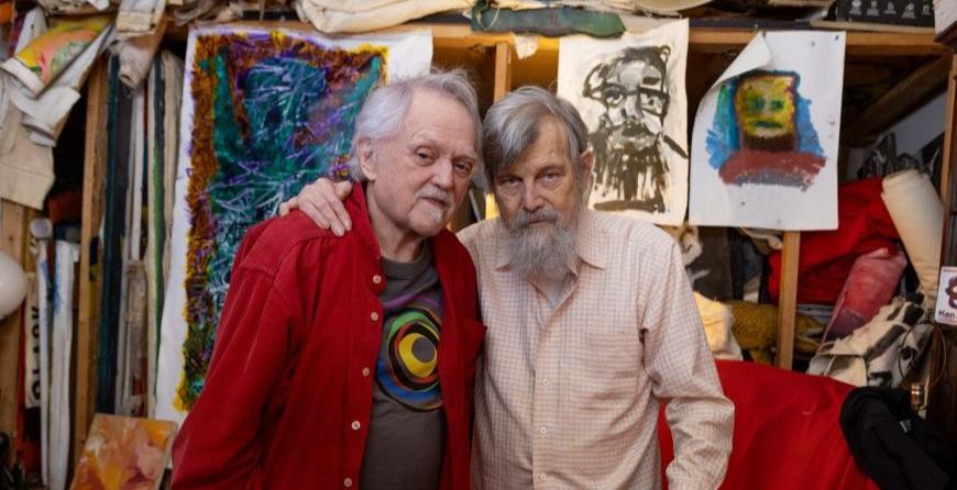 Joel and Jim stand together in their living room, arms around one another. 