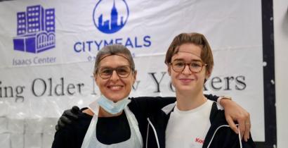 Two kitchen volunteers, dressed in aprons, smile at the camera. 