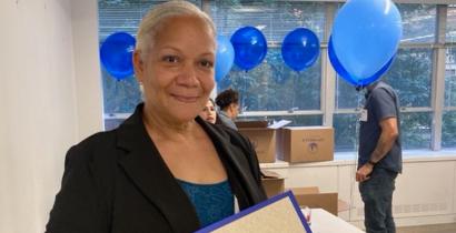 An older woman with light brown skin and grey hair holding an award in front of blue balloons.